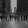 A black and white photo of about 10 people linked arm-in-arm across a city street. The street is slick with a recent rain, and some people in the photo are wearing rain gear and ponchos. The city rises around them with street lights and tall buildings in the background.