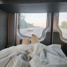 Two legs in thick casts under a blanket are seen from the inside of a hospital van. The highway stretches out beyond the back windows.