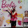 A girl in a red shirt with a reindeer hat, scarf, and tutu stands in front of a Barbie poster.
