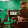 Film set with a director and two cameramen visible, filming a couple in period costumes against a large green screen