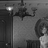 A black and white still from the 1944 film Gaslight. Paula, played by Ingrid Bergman, stares up with a haunted expression at a flickering gaslight chandelier in her home. Just one of its light is on.