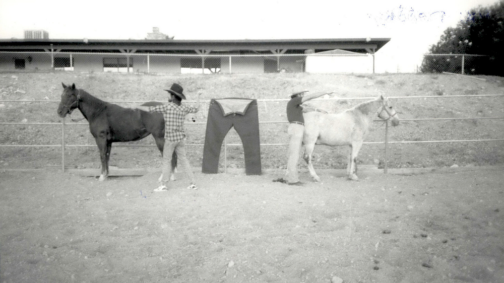 A black and white photograph of two horses standing in front of a fence and facing away from each other. The horse on the left is colored black, and the one on the right is colored white. They are pulling on a pair of trousers hanging between them, with two men standing next to each of them in the foreground, pointing in opposite directions. There is a long horizontal building with a roof a few feet in the background, at a higher elevation than the horses.