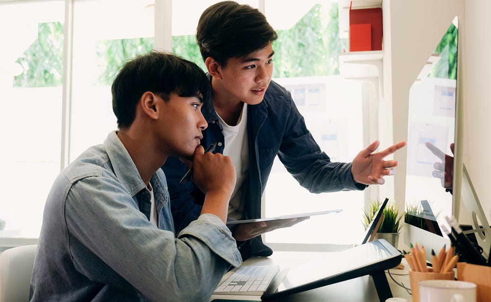 Two young men review a design on a large iMac computer monitor in a bright, sunny room.