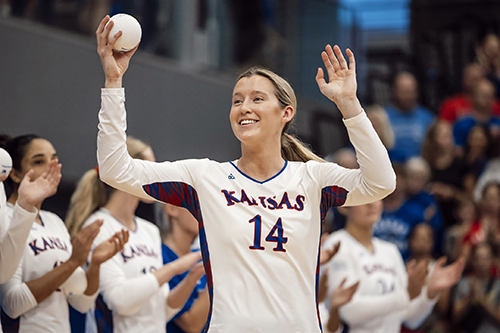 A volleyball player waves to the crowd while holding a small volleyball in her hand. 