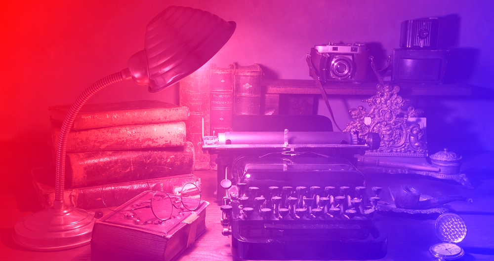 The image shows an old typewriter on the table, illuminated by a lampshade. The image also includes several books on the table, a camera, a pair of glasses on top of a book, and a pocket watch on the table.