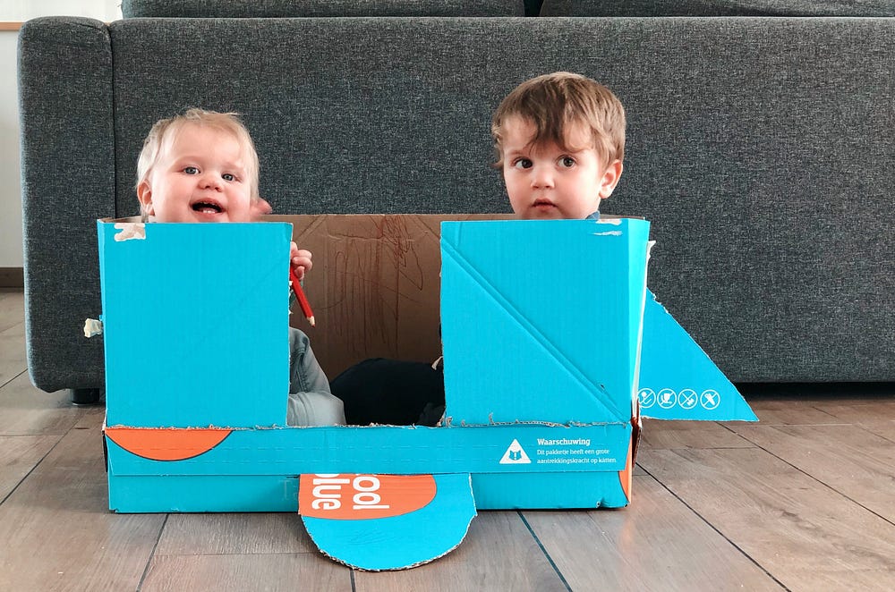 Two toddlers sitting in a cardboard box, cut into the shape of a rocket ship.