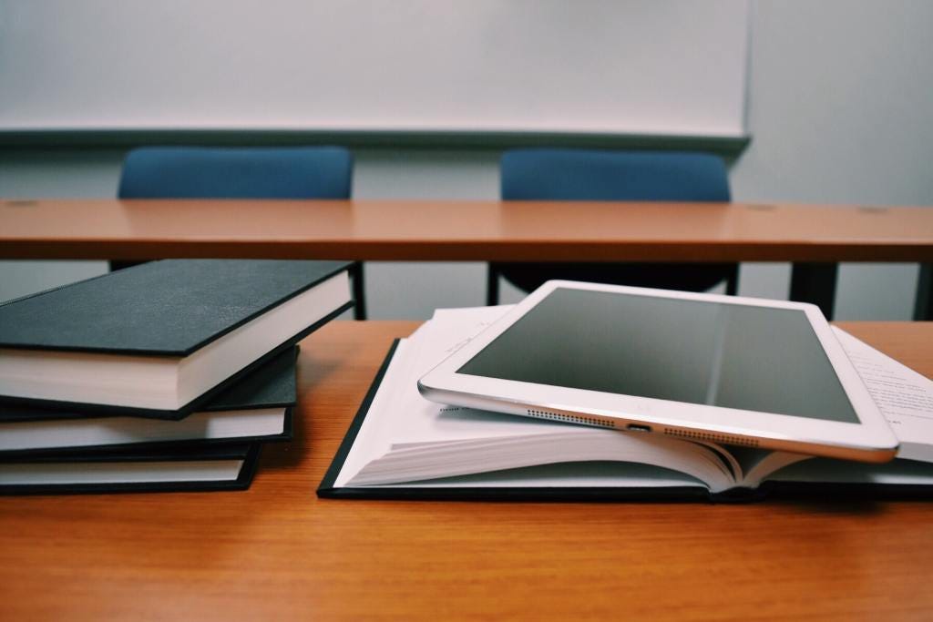 A classroom table with books and a tablet device ready to be used for studying, and there is a whiteboard in the background. Pexels.com Pixabay