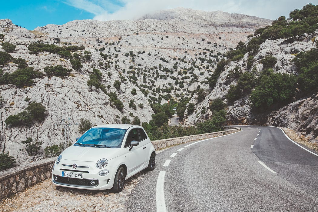 Imagen de un automóvil Fiat 500 blanco estacionado al lado de una carretera sinuosa. La carretera está rodeada de un terreno montañoso rocoso con vegetación escasa. El cielo está parcialmente nublado y el entorno parece estar en una ubicación natural, posiblemente remota. 