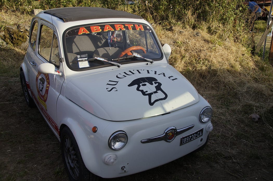 Imagen de un pequeño automóvil Fiat vintage con modificaciones. El coche es predominantemente blanco con marcas y logotipos, incluyendo “ABARTH” en el parabrisas y un logotipo de escorpión en el capó. También hay otros textos y números como “SU CINTA” encima del logotipo del escorpión y una matrícula que lee “0927 CCJ”. El vehículo tiene características de estilo de carreras como una jaula antivuelco visible en el interior. Esta imagen podría ser interesante o relevante para aquellos que aprecian los coches clásicos, la historia automotriz o las modificaciones de carreras.