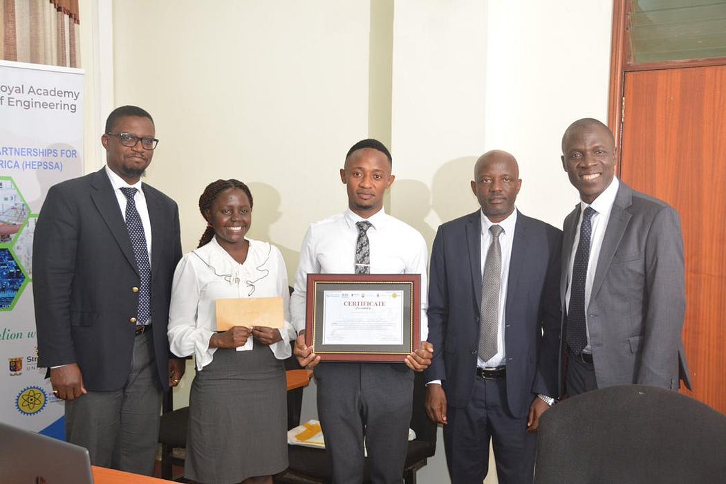 Elizabeth and Henry stand with a group of smartly-dressed officials, smiling with their certificates.