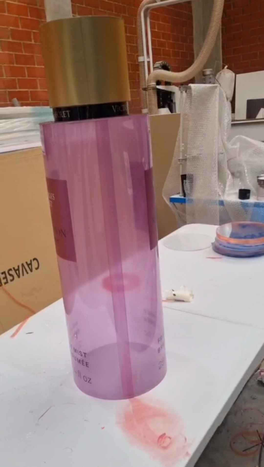 Cylindrical translucent pink and purple mica perfume display dummies with matte gold caps and central pink labels shown inside a workshop.