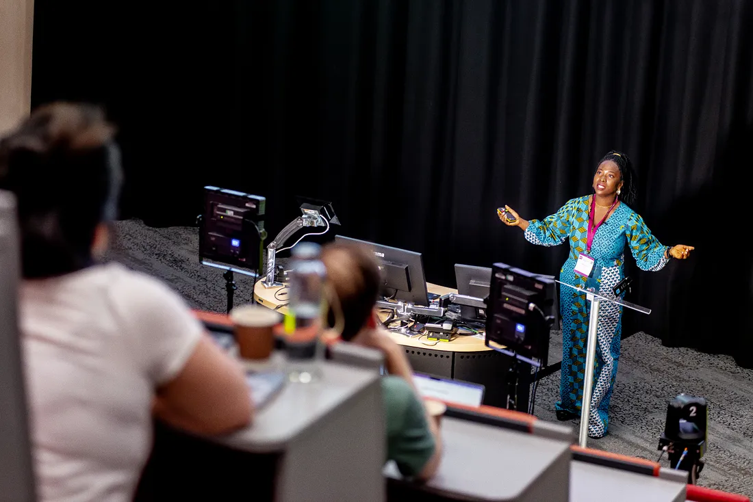 A full length image of the same African female, Debs, standing behind a lectern, with arms spread out beside her, as she speaks to an attentive audience.