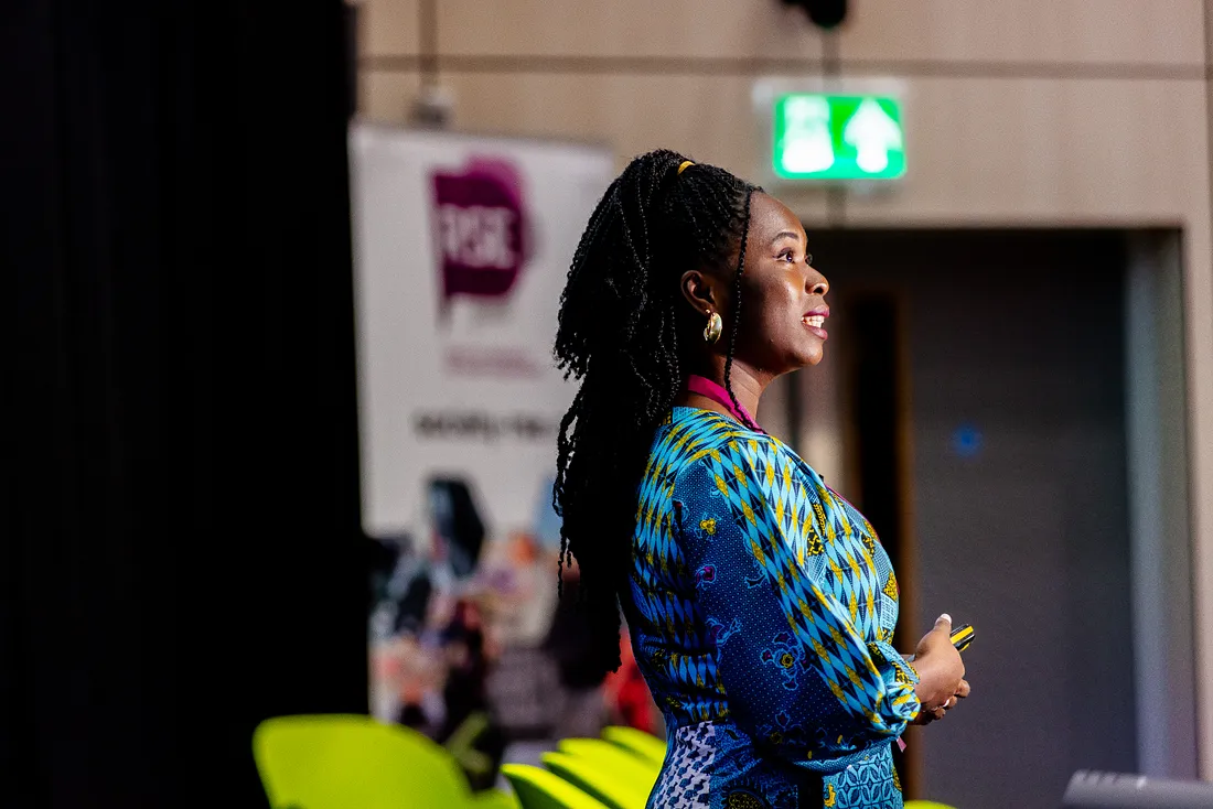 An image of a speaker, Debs, delivering a presentation. Debs is a black female, dressed in a blue and yellow jumpsuit made from African "ankara" prints. Picture shows her side profile, with an RSE banner blurred out in the background.