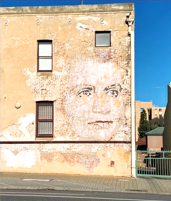 호텔 벽에 있는 거대하고 희미해진 여성 얼굴 초상화의 거리 풍경. 옅은 노란색 벽돌로 둘러싸여 있다.<BR>