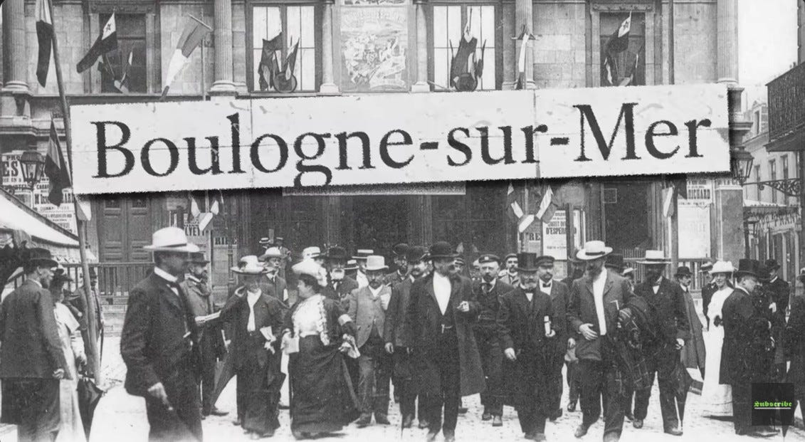Black‑and‑white photograph of delegates at the first Esperanto Congress in Boulogne‑sur‑Mer, France, 1905. Men and women in formal attire walk past a building decorated with flags, marking the birth of Esperanto’s international movement. Photo from Wikipedia, licensed under CC BY‑SA 4.0.