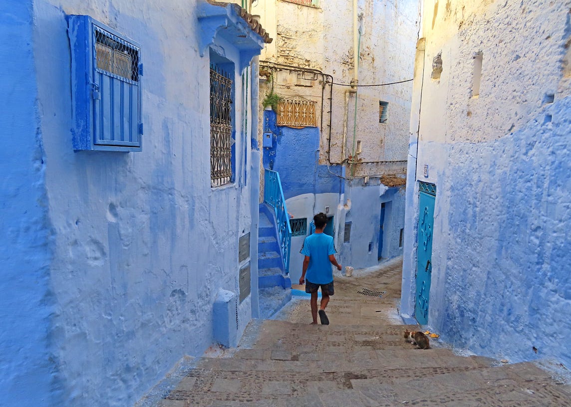 Man in a blue tshirt walks down a narrow street with blue, plastered buildings on either side. Blue doors, blue windows, blue stairs.