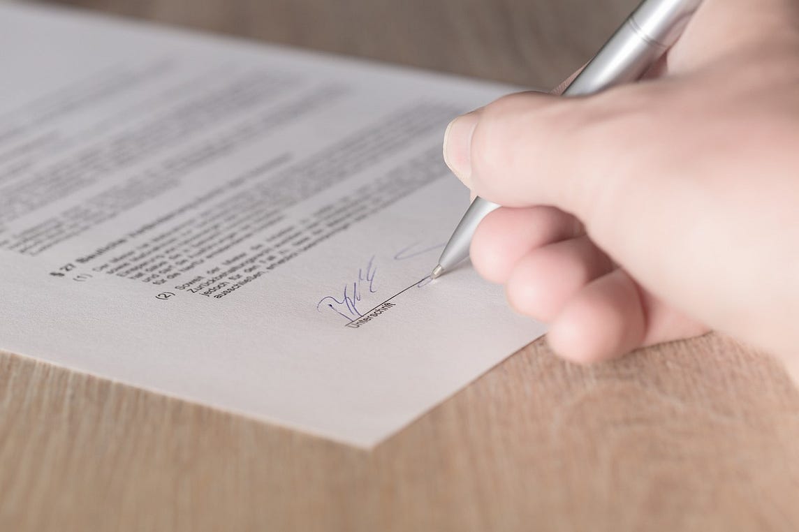 IMAGE: A person’s hand with a pen signing a contract