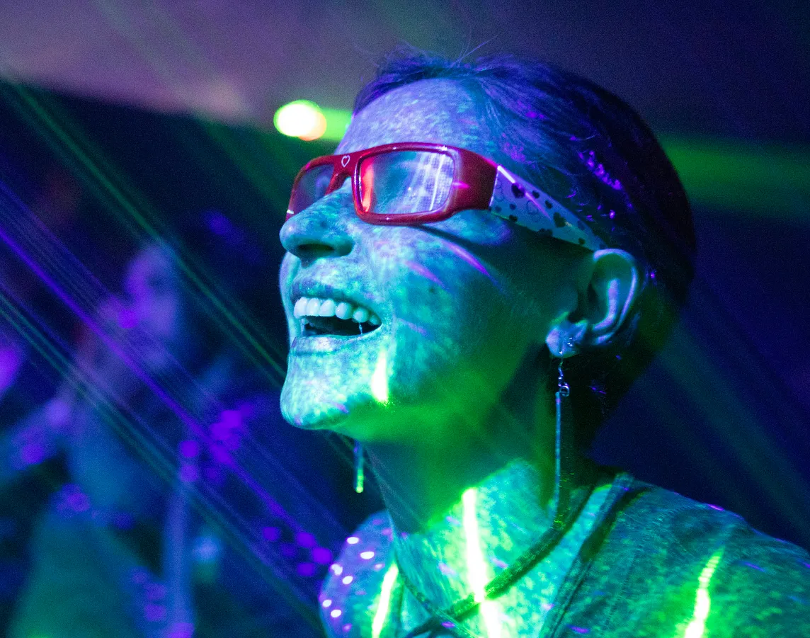 This image depicts a person at what seems to be a party or event where UV lighting is in use, as indicated by the vibrant greenish glow on their face and clothing. The person is wearing red-framed glasses and earrings, and appears to be having a joyful moment, with their mouth open as if they are laughing or cheering. The background is blurred, but lights and possibly other people can be seen, suggesting a lively atmosphere. The visible laser lights add to the festive ambiance of the setting.