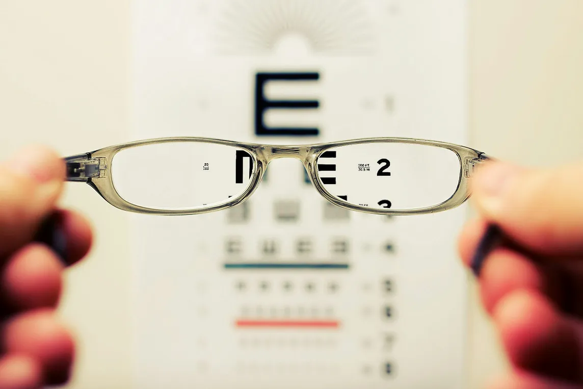 An eye chart as seen through the lenses of eyeglasses held by two hands.