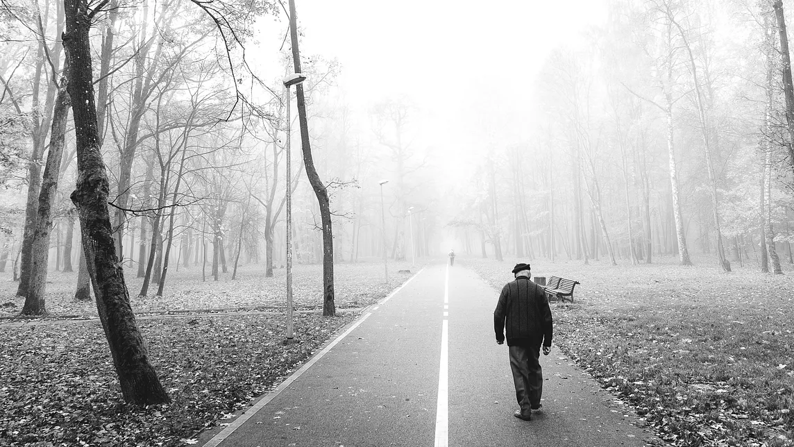 In a moody black-and-white photo, an old man shuffles along a lonely road.