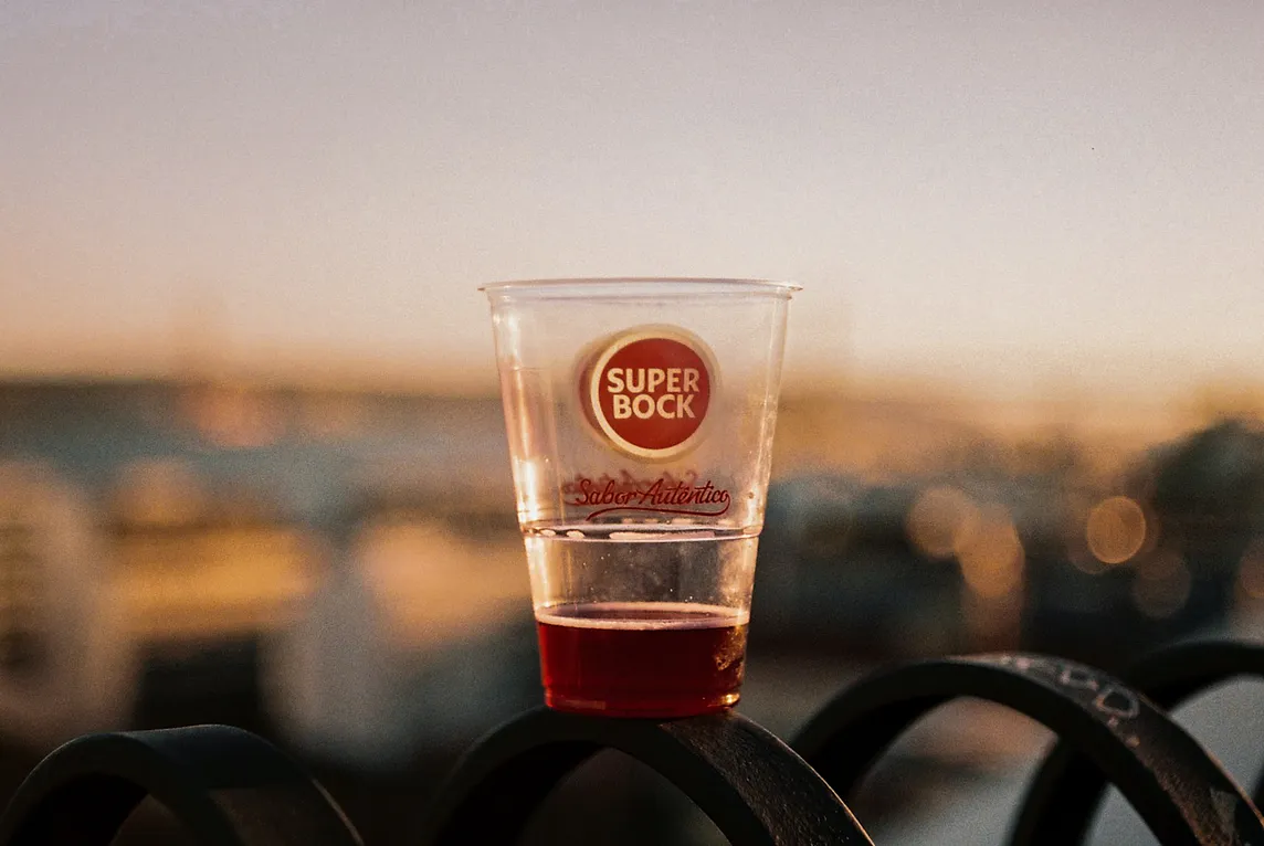 A half empty plastic cup of Portuguese beer sitting on a fence in evening light