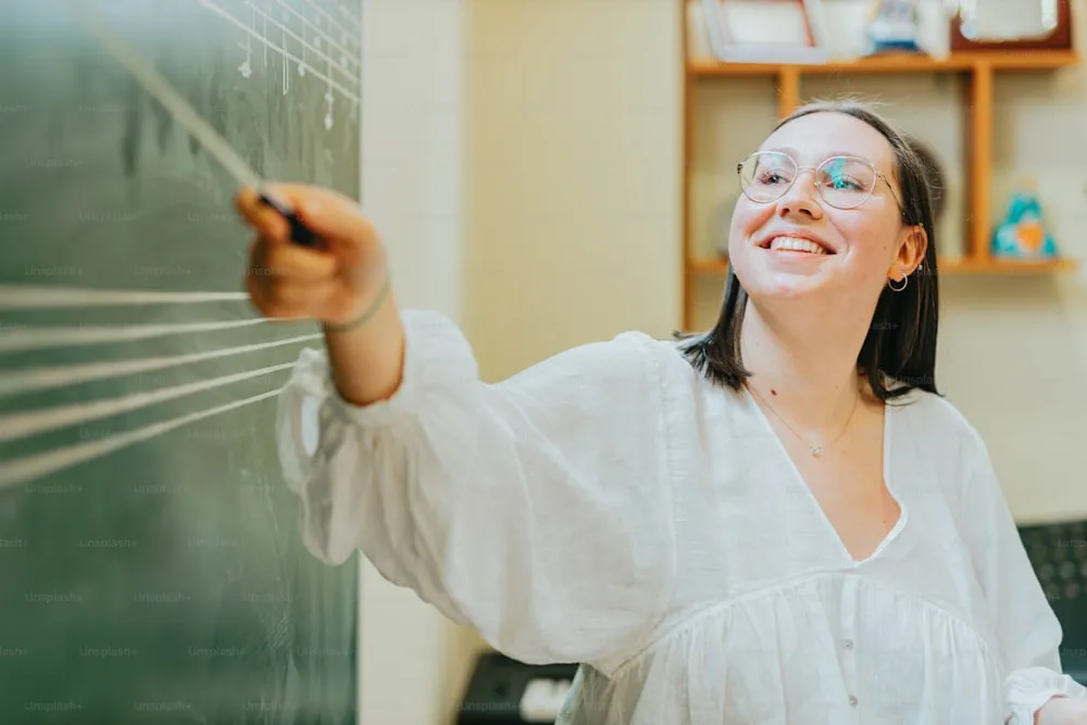 A young and happy music teacher stands in front of an old fashioned chalk board with the musical staff and some musical notes written on it. She is pointing to the music with a conductor’s baton. She is wearing classes and a white, flowing blouse. She seems happy to be teaching.