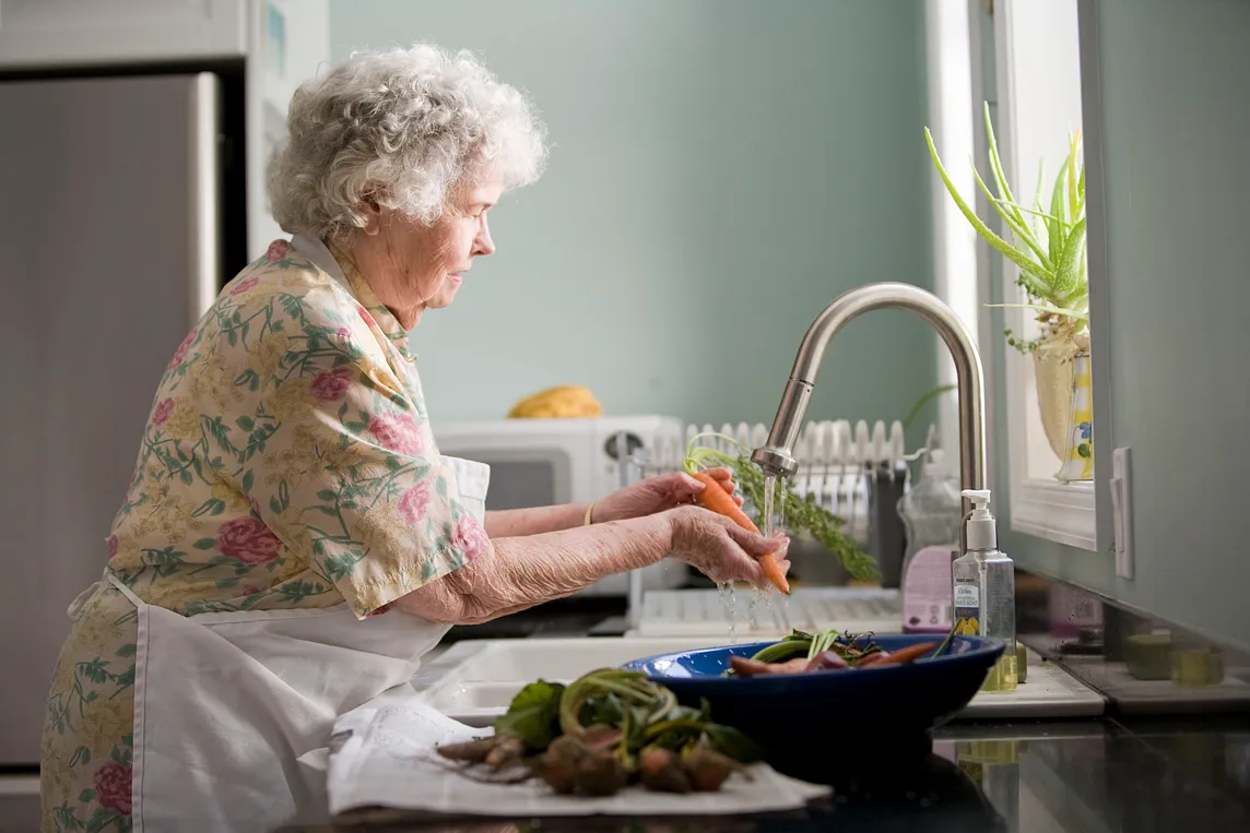 An older woman stands at the sink, rinsing a carrot.