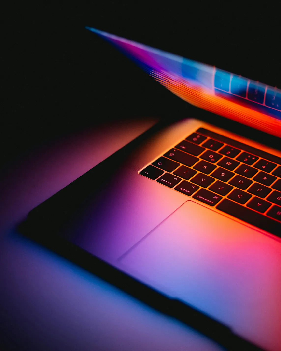 Close-up view of a backlit laptop keyboard in dark setting, illuminated with dramatic gradient lighting in purple, orange and blue tones, creating a moody tech aesthetic