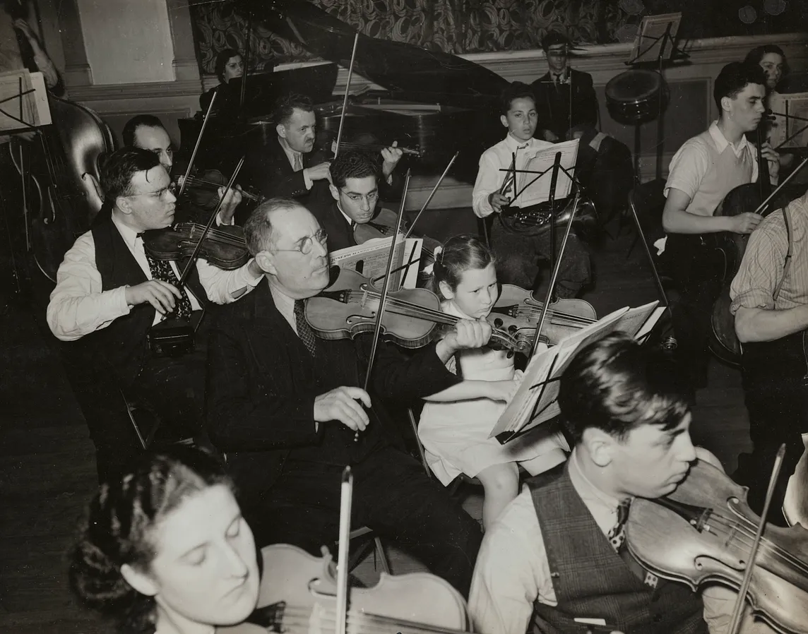 a portion of an orchestra of both adults and children mostly playing violins with a piano in the background.