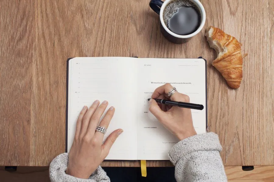 Woman’s hands on a paper journal as she is writing goals for a trading journal. Paper journal is on a table with a bitten-into croissant and a cup of coffee nearby.