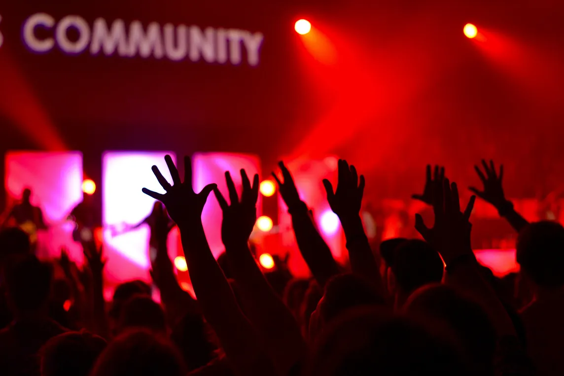 Hands raised at a live show with “Community” appearing on the screen on the stage.