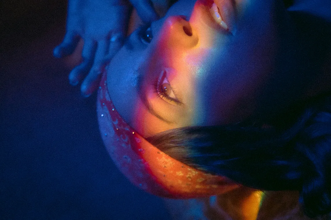 A blue toned image of a woman looking away. She wears a orange bandana and rainbow of light glows across her face.