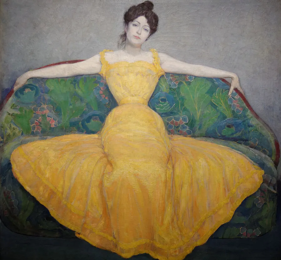 “Woman in a Yellow Dress” (1899) by Max Kurzweil, via Wikimedia Commons. A painting of a woman with an hourglass figure in a yellow gown, reclining on a floral-patterned sofa. Facing the viewer, she has her arms draped over the backrest, exuding a relaxed yet confident attitude, with a hint of collected impatience on her face.