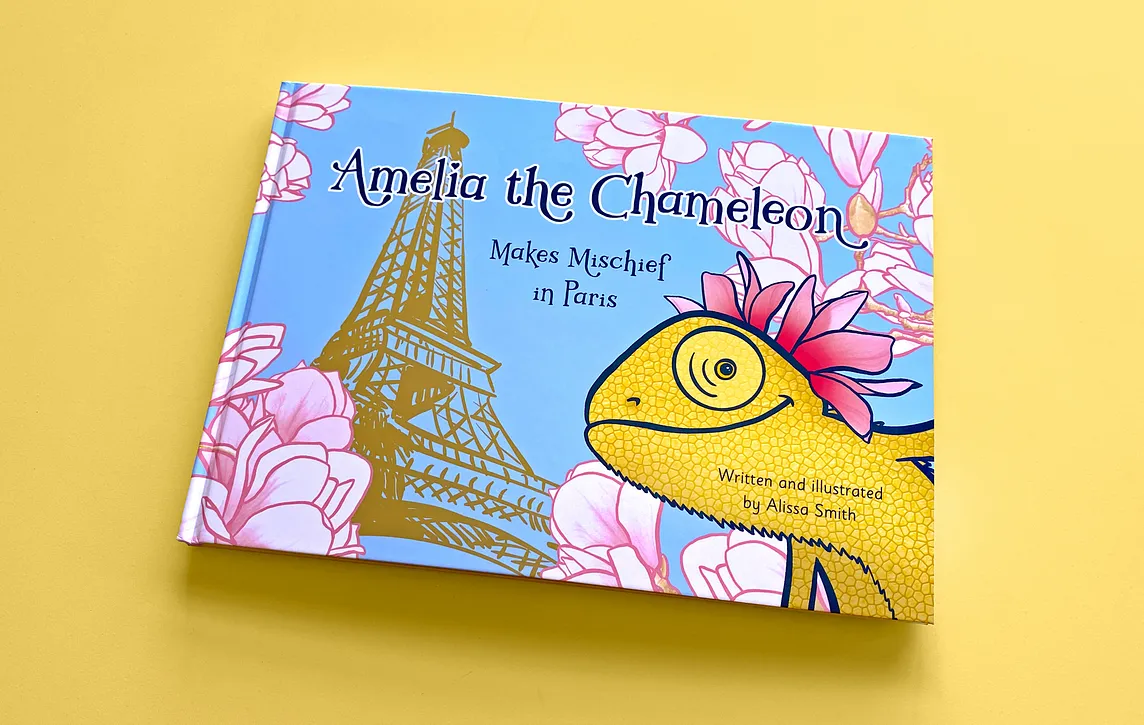 A hardcover children’s book called “Amelia the Chameleon”, subtitled “Makes Mischief in Paris”. An illustrated of a gold chameleon with magnolia flowers on its head smiling in front of a sketch of the Eiffel Tower on a blue background. Magnolia flowers frame the edges. The book sits on a yellow background.