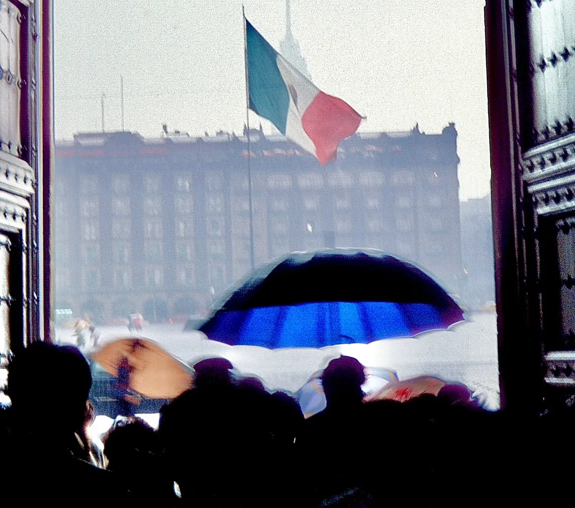 A Mexican flag blows in the wind in a large open square during a rainstorm, photo is taken through large wood antique doors of the National Palace.