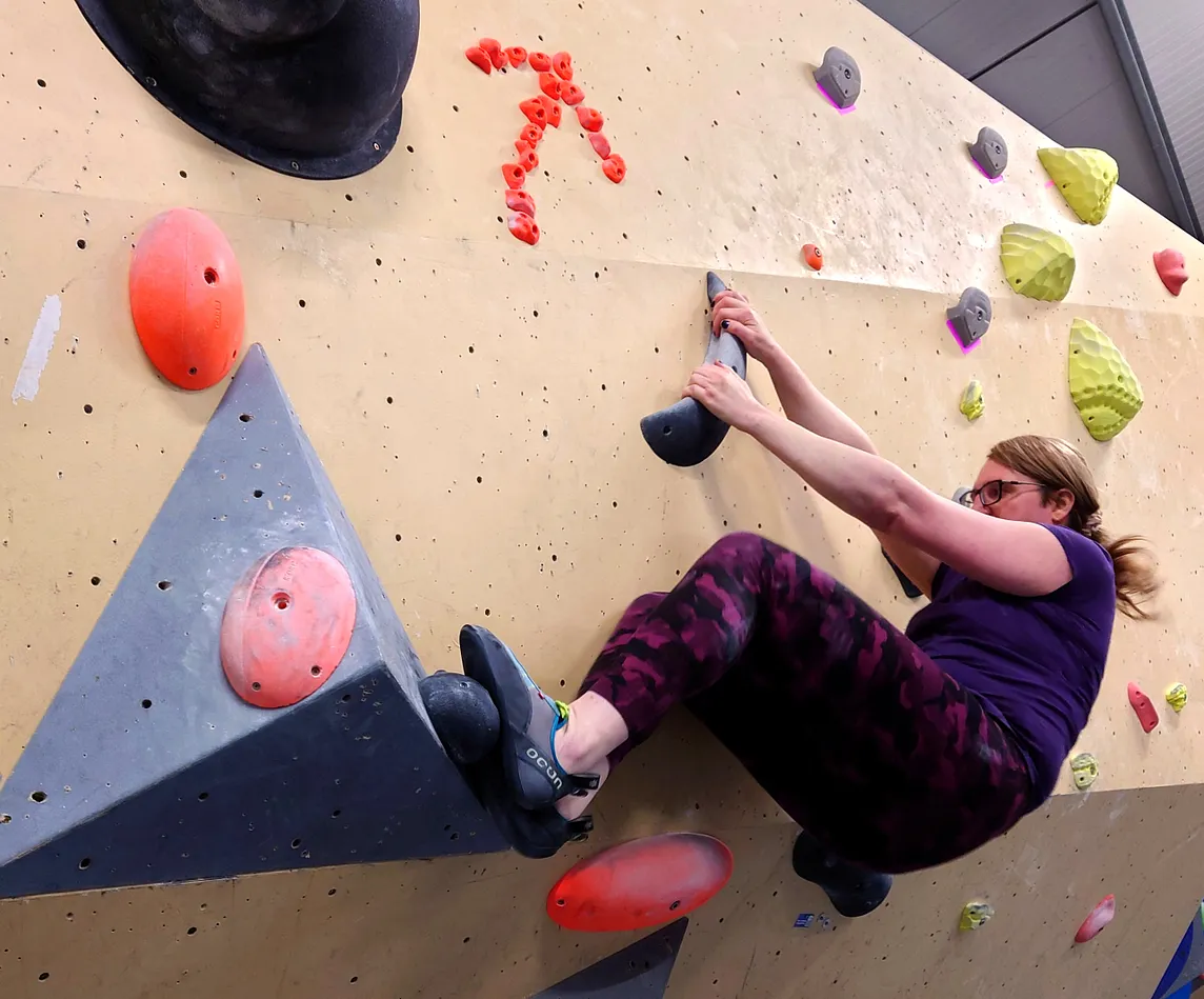 The author on a bouldering wall in layback position, hair tied back, wearing purple camo leggings and a violet tee shirt. Above them is an arrow pointing upwards, made out of tiny footholds.