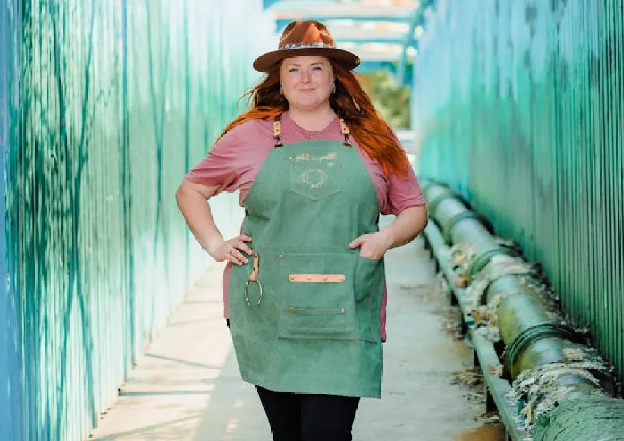 A woman with red long hair wearing a hat, green apron, and a smile.