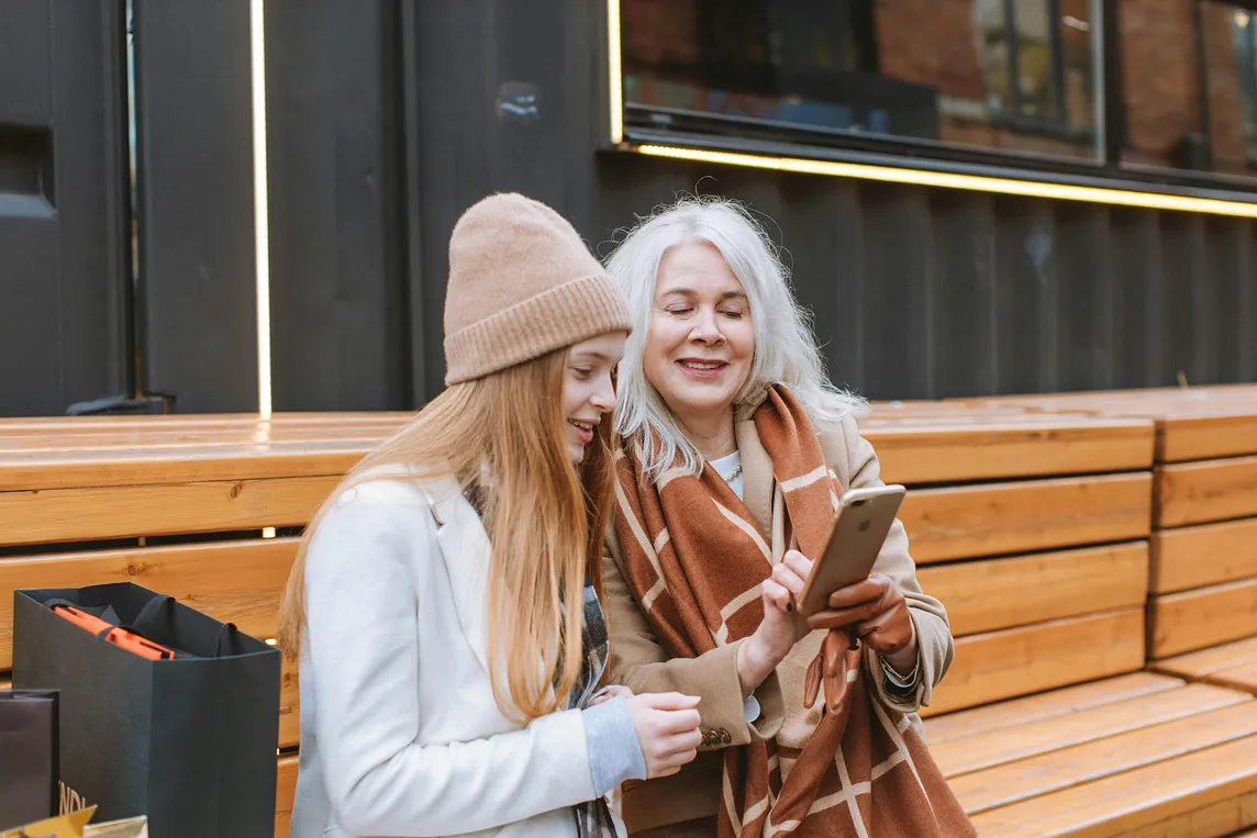 An elderly women sitting on a bench next to a much younger women as they both look at a phone.