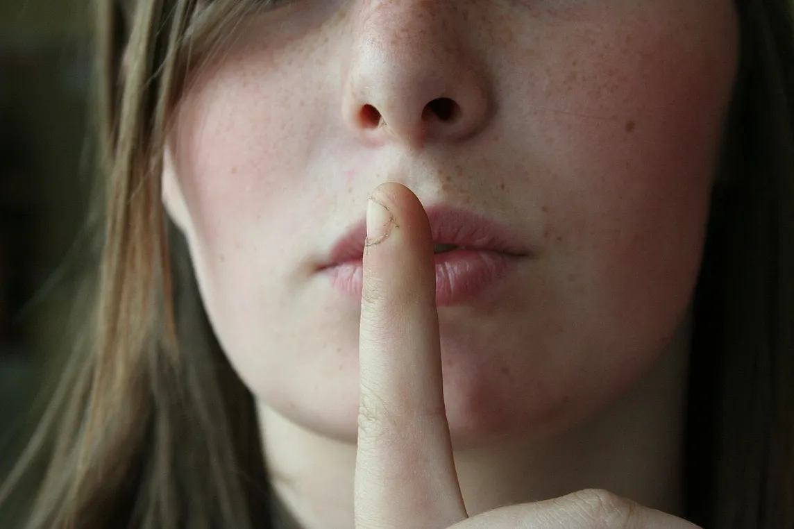 Image shows part of a face where a finger is hold in front of the lips to hush others.