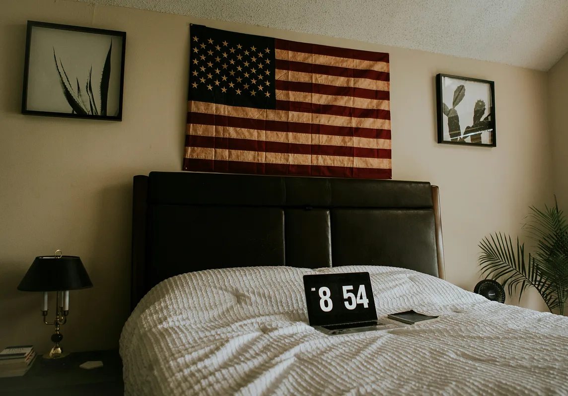 flag above bed headboard, laptop on bedsheets showing 8:45