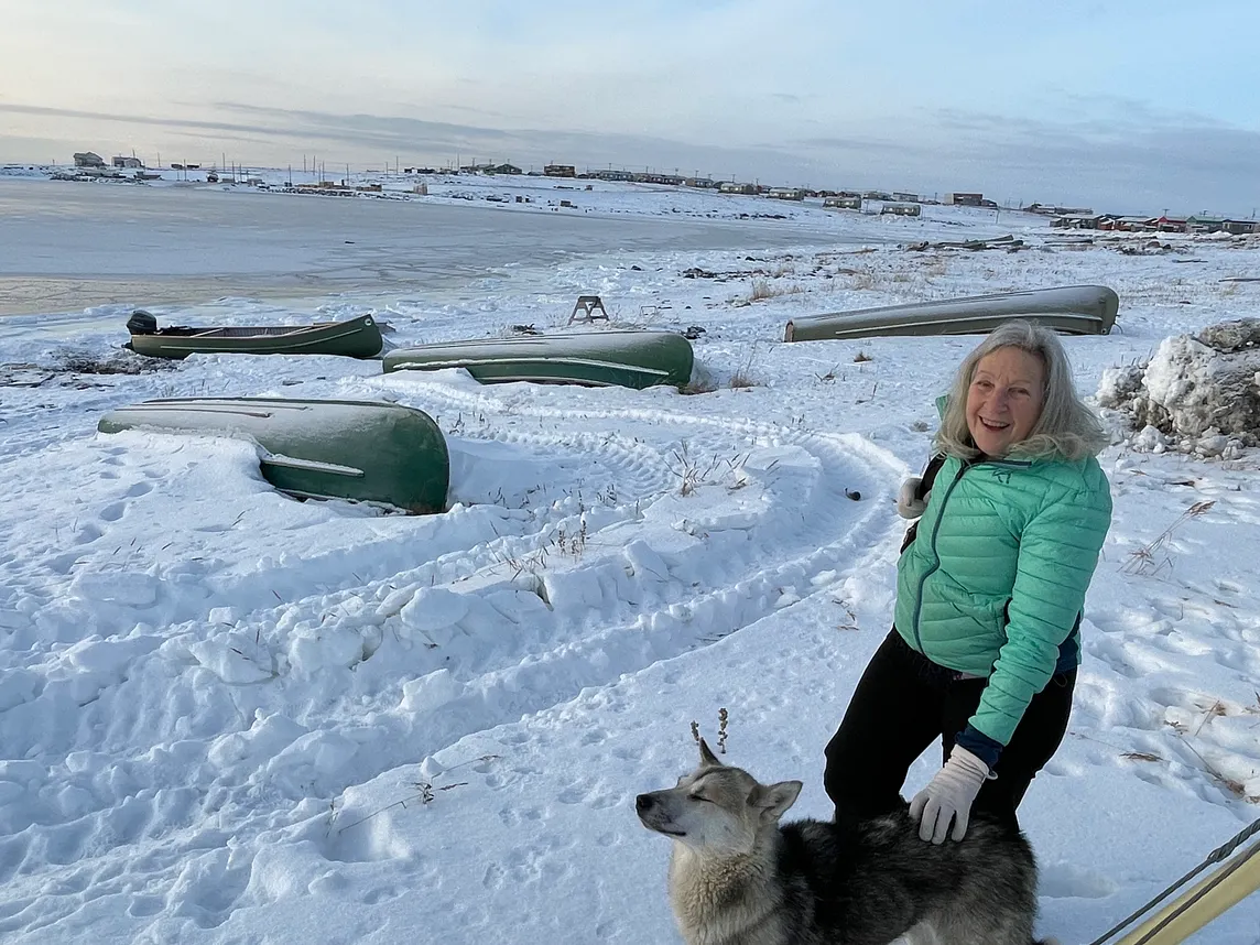 Woman wearing a green puffer jacket, smiles into the camera while petting a dog. They are on the snow covered shore of a body of water. Four overturned boats are on the snowy beach.
