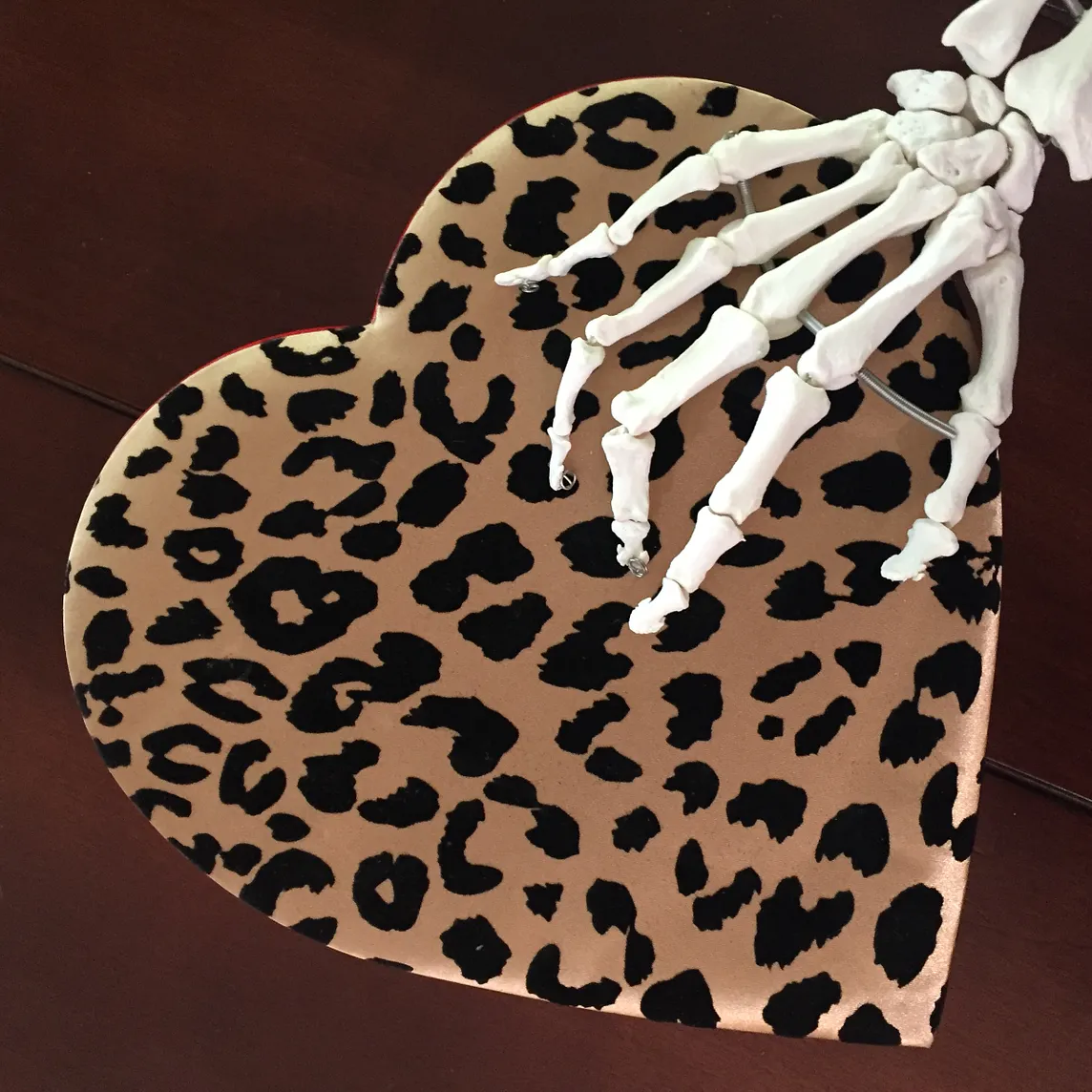 A skeletal hand clutches a heart-shaped box covered in leopard skin-patterned velvet.