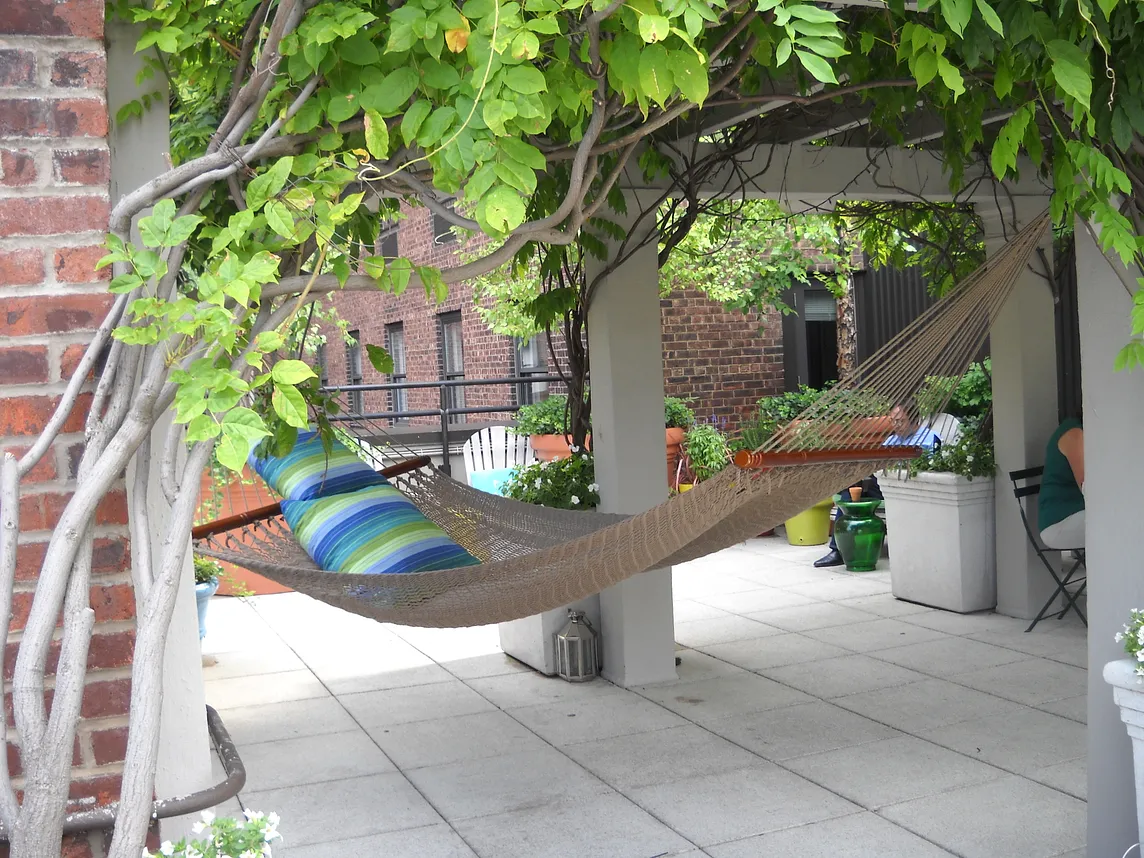 A hammock with a blue and green striped pillow hanging on an exterior patio, with tree leaves above the hammock, a brick multistory building in the background.