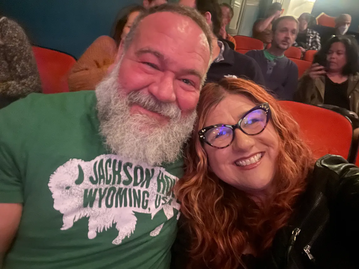 Two 50-something best friends — a bear of a guy and his bespectble BFF — in theater seats, smiling.