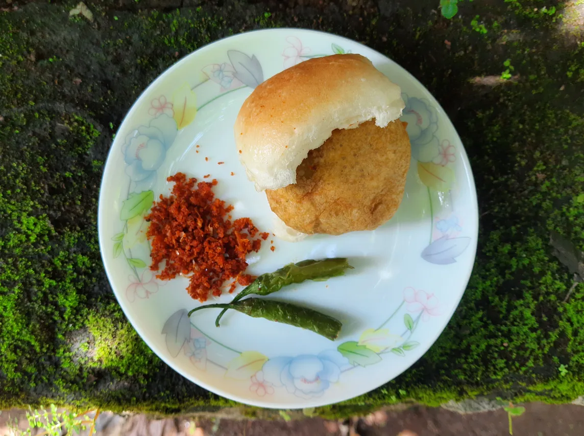 In a plate, a potato fritter is sandwiched between a bread bun with two green chilies and a dry chutney served along with it.