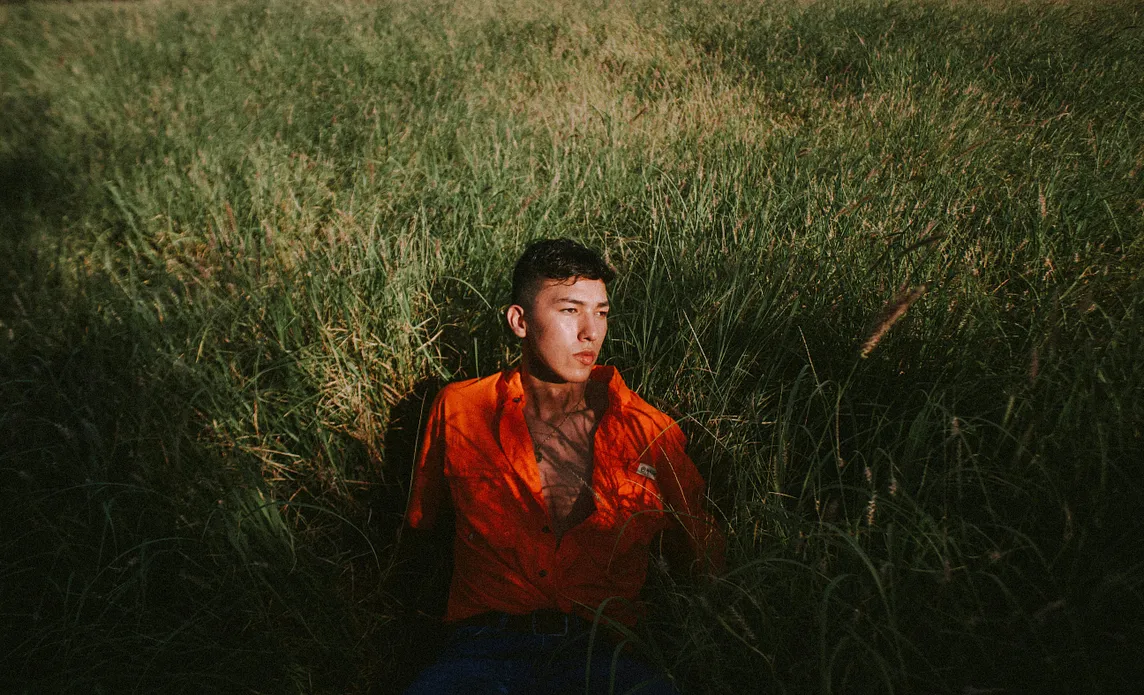 A young man in a field of tall grass, half sitting up and looking off to the side.