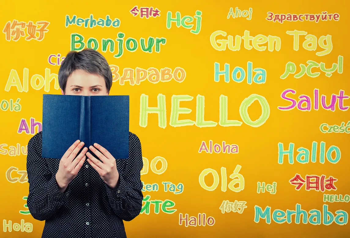 A student woman holding a textbook over a yellow wall written with the word hello translated in different languages.⁠