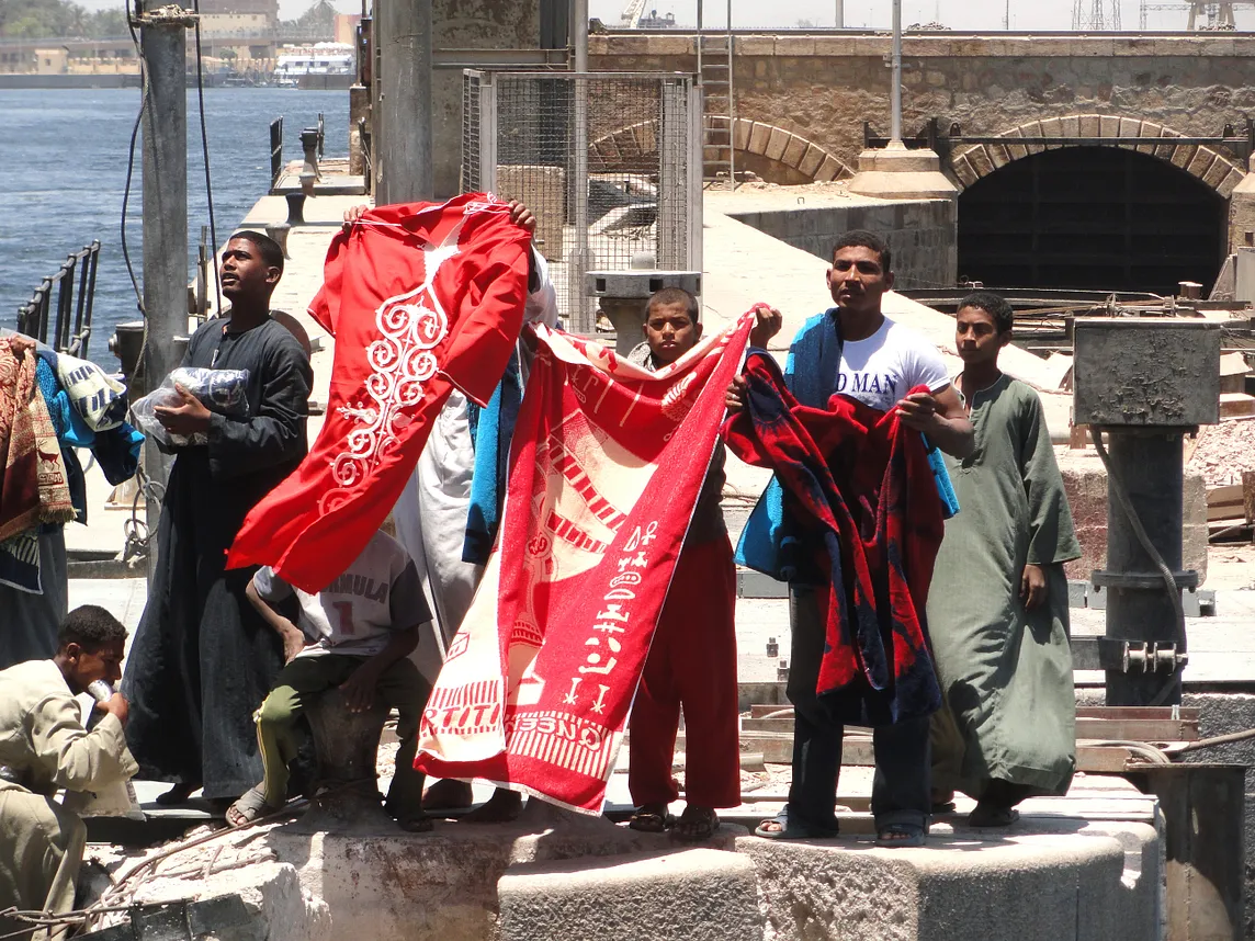 Men on the dock beside a river holding up blankets and other goods to entice cruise passengers