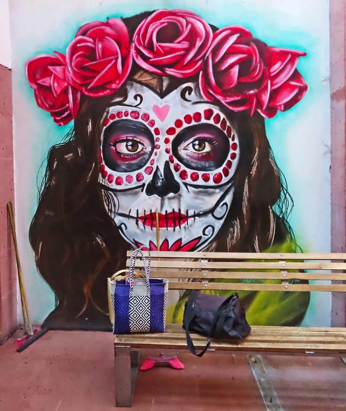 Streetart painting of a woman Catrina — her face painted up to look like stylized skeleton with red lips and red dots around the black rings of eyes and a red flower design on her chin. She has a crown of red roses in her hair. There’s a wooden bench in front of the streetart that holds a blue bag and a purse. Underneath is a pair of pink slip-on shoes.