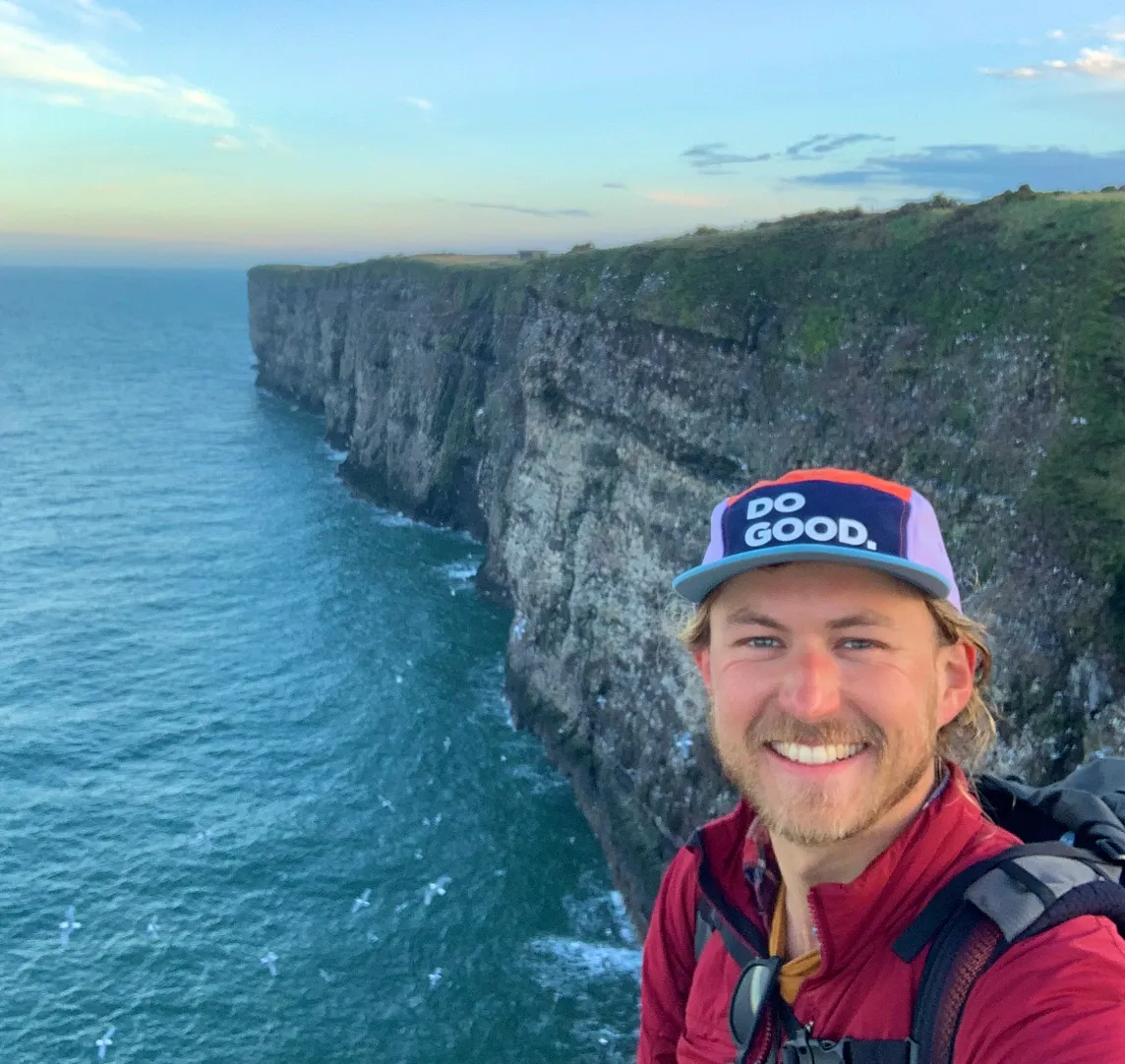 A hiker smiles happy with sea cliffs in the background.